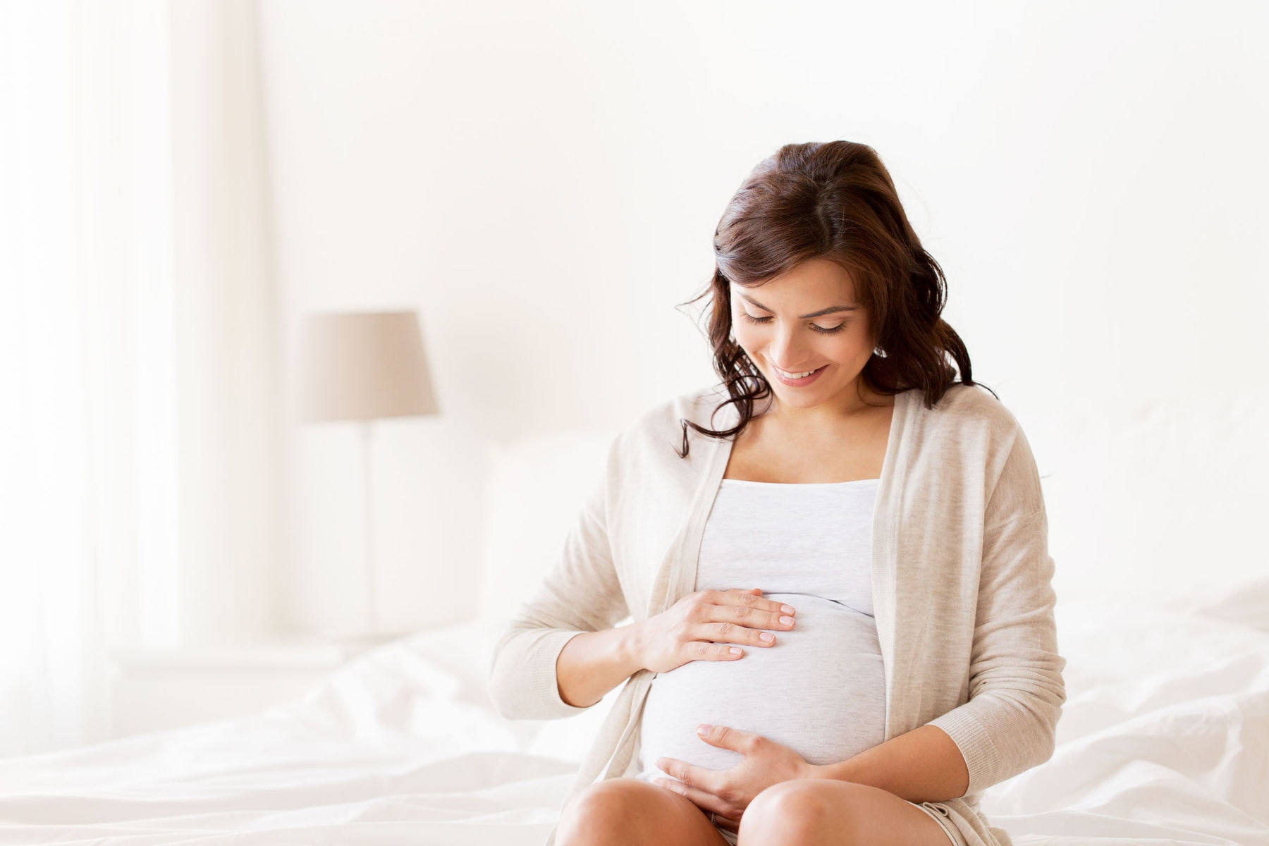 pregnancy, rest, people and expectation concept - happy pregnant woman sitting on bed and touching her belly at home